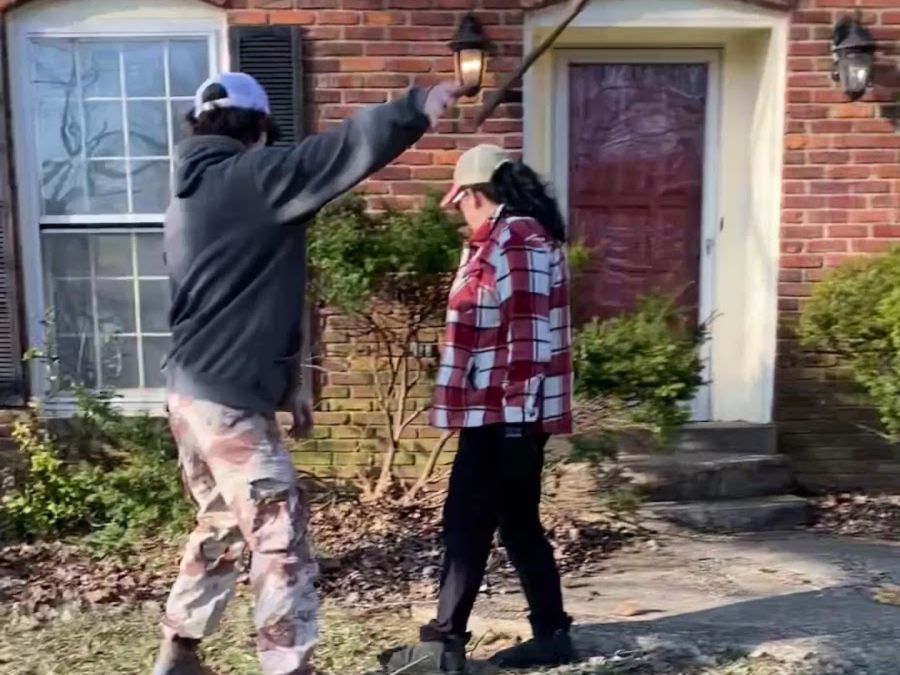 Two people working on a front garden at a red brick house in early spring, one reaching up to cut back a long branch and the other clearing the bed below, with dead grass and leaves raked across the lawn.